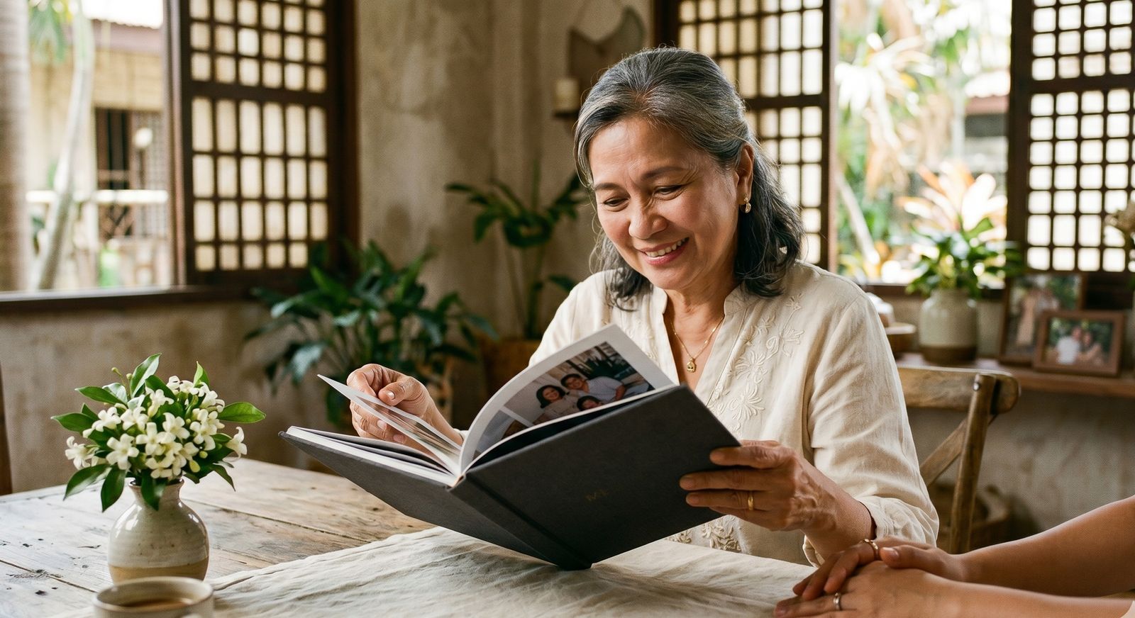 Filipino mother in her late 50s opening a hardcover photo book at a sunlit dining table with her adult daughter beside her